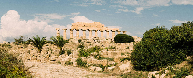 The ruins of a columned stone building in a rocky setting, with a blue sky in the background